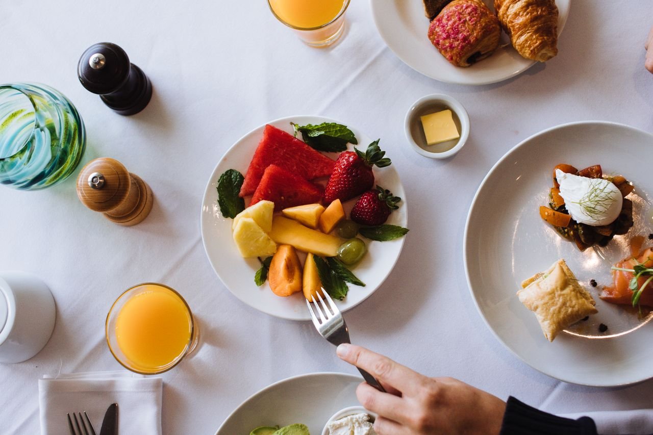 Top view of crop person eating fresh delicious cut fruits on table with white tablecloth during breakfast
